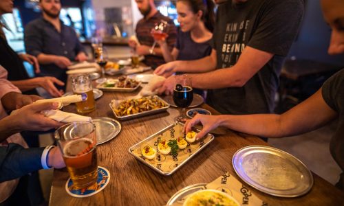adults gathered around table at Cob & Pen in Lakeland, FL drinking beer and sharing small bite plates of deviled eggs, dip, french fries and wings
