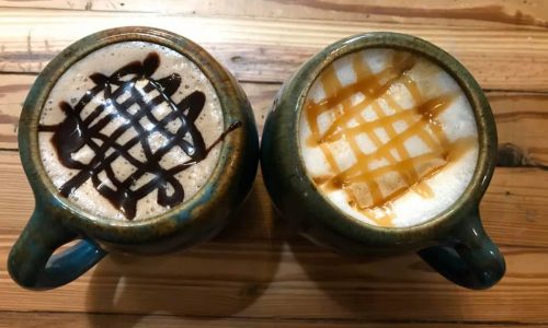Two cups of coffee on wooden table. One with chocolate drizzle and one with caramel drizzle.